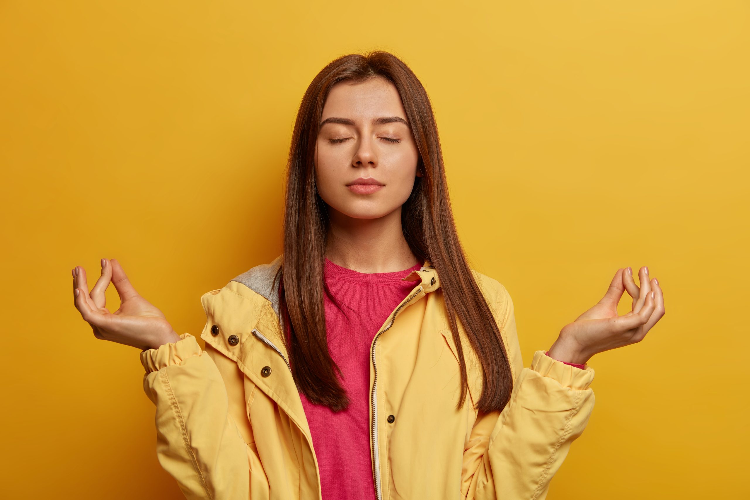 Relaxed dark haired young woman with healthy skin, makes zen mudra gesture, tries to calm down after hard day, breathes deeply and practices yoga, wears windbreaker, isolated on yellow background